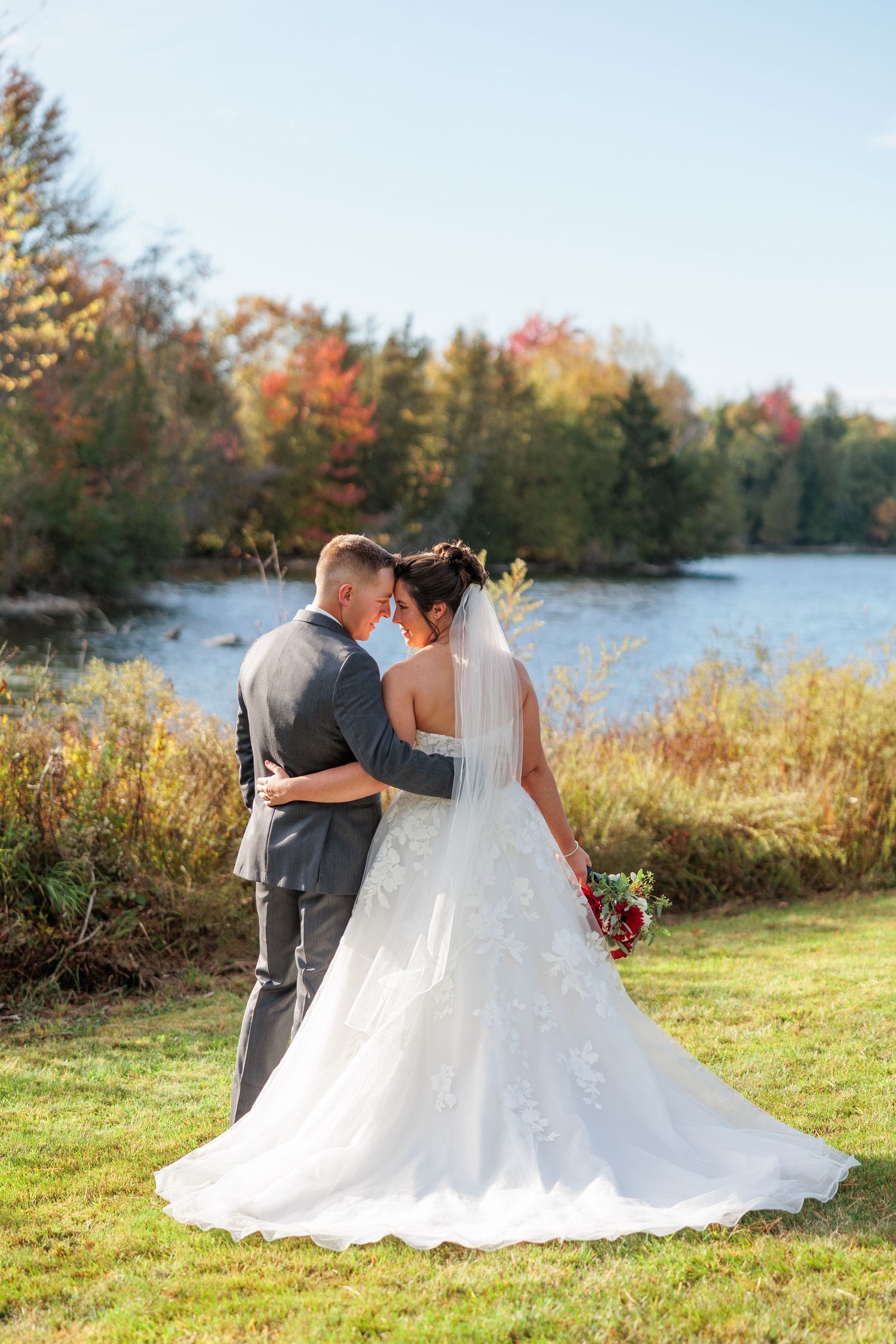 Bride and groom embracing near a lake surrounded by vibrant fall foliage on wedding day.