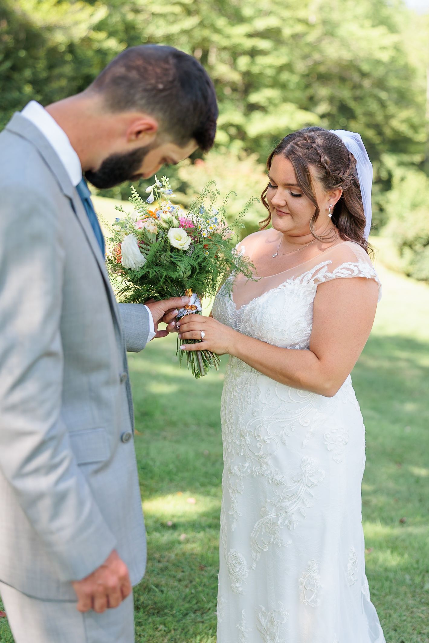 Bride in lace gown smiles at colorful bouquet during first look with groom.