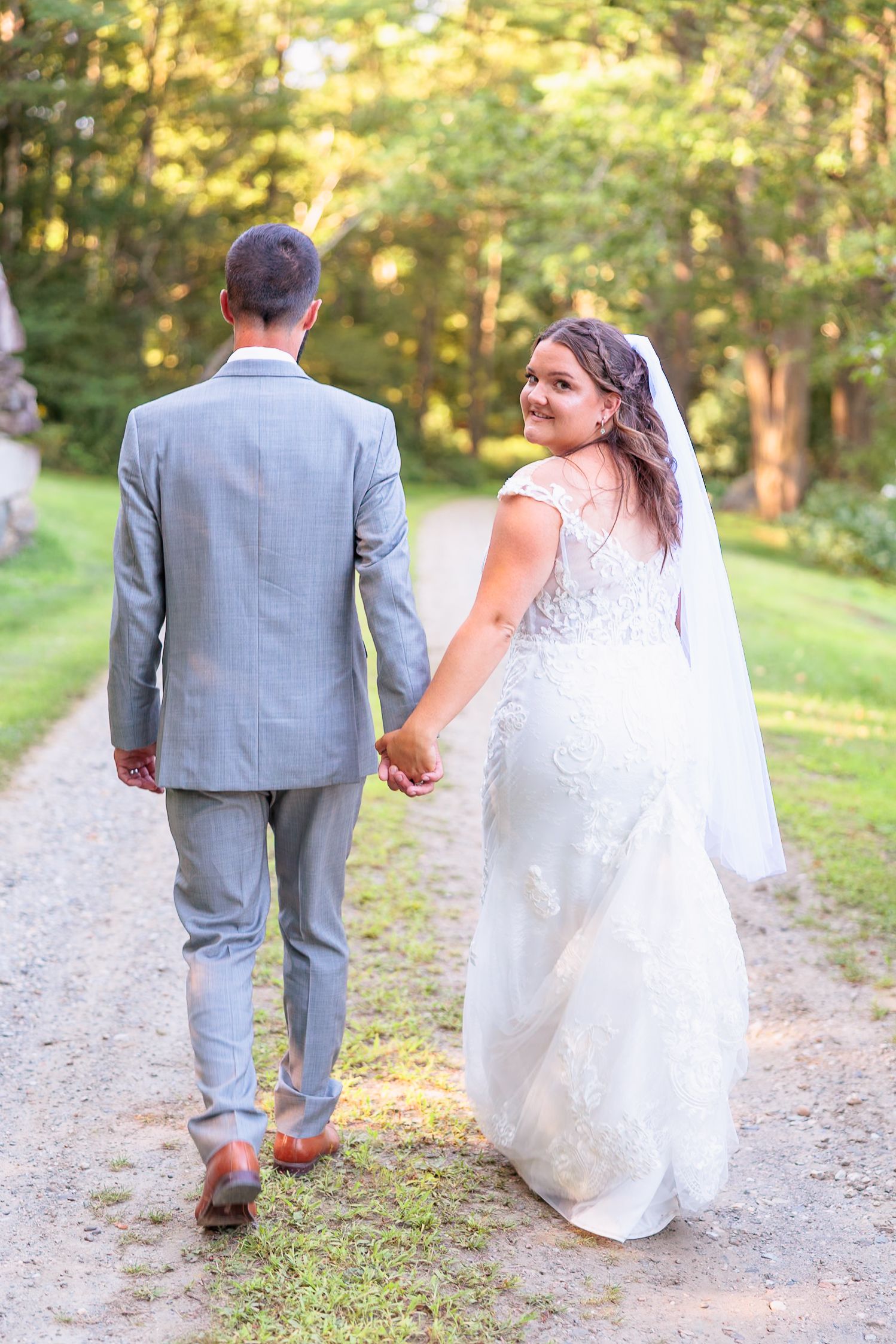 Bride in lace gown glances back smiling while holding hands with groom walking down a sunlit garden path.