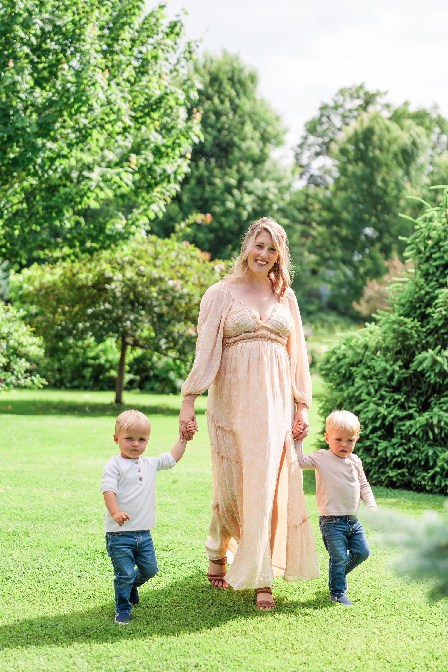 A smiling mother in a peach dress walks with her two toddler sons through a lush green garden.