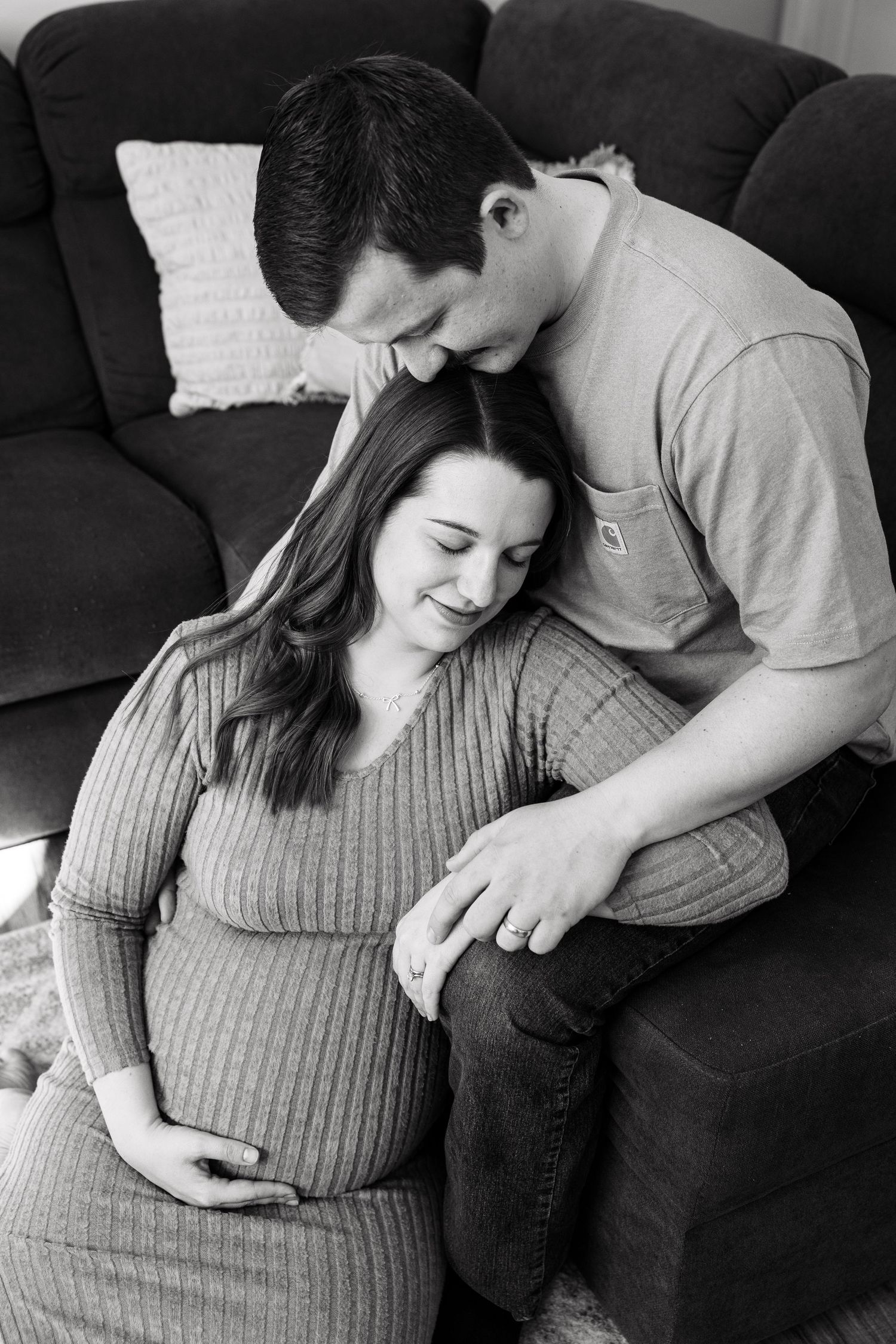 Black and white photo of a couple sitting by a couch, man kissing woman's head as she cradles her bump.