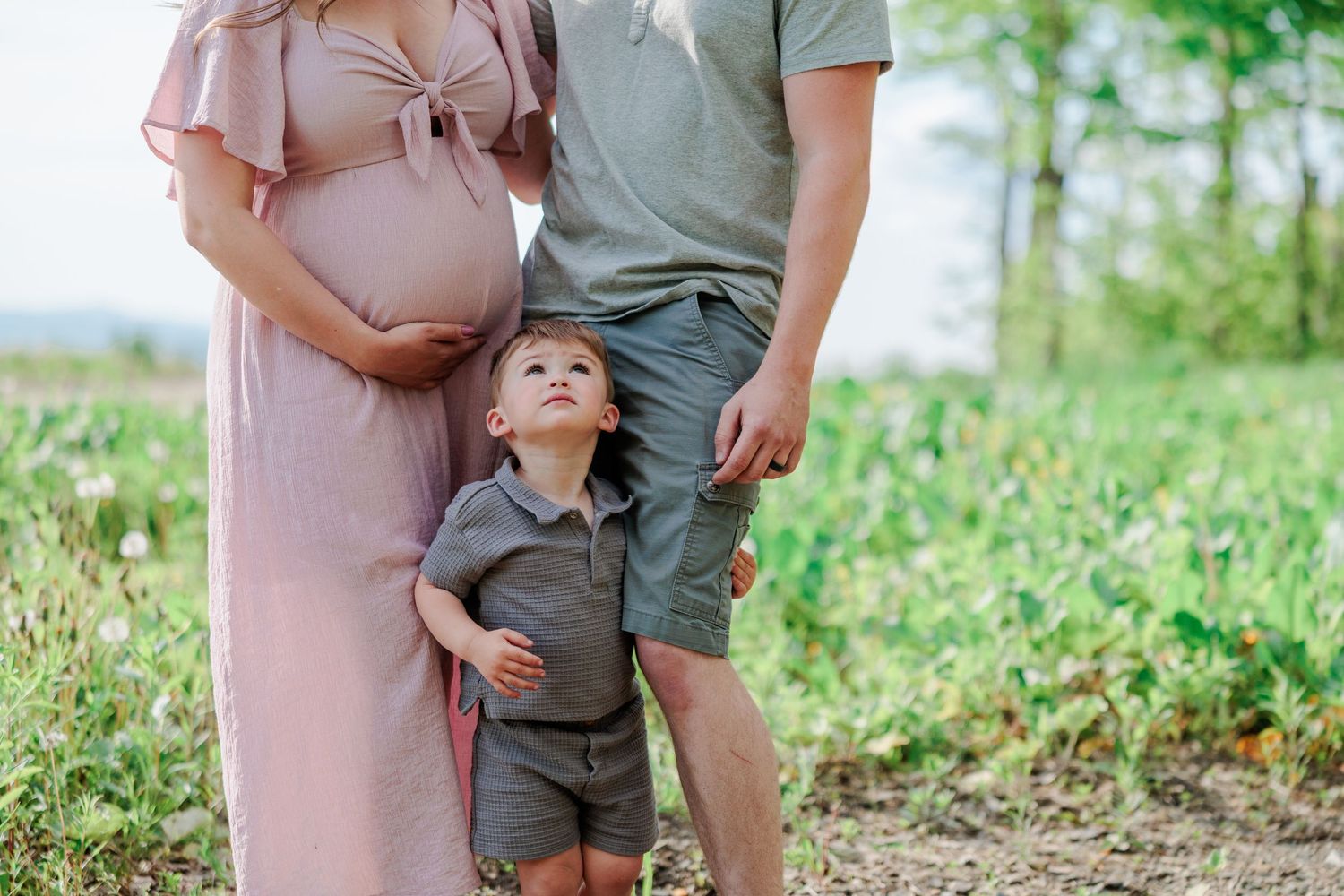 Pregnant woman and man stand with toddler boy between them outdoors.