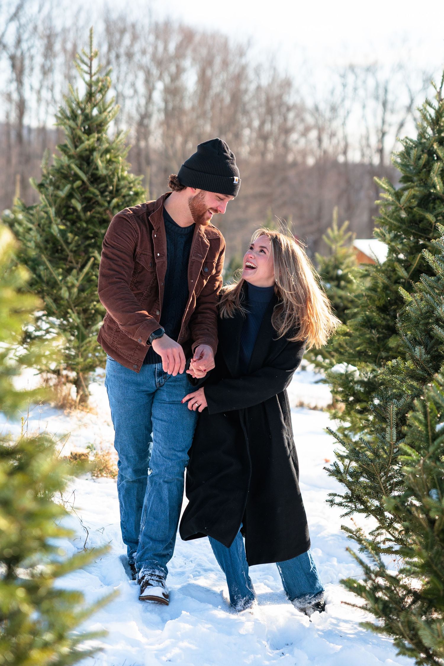 Happy couple holding hands and laughing while walking through a sunny snowy Christmas tree farm.