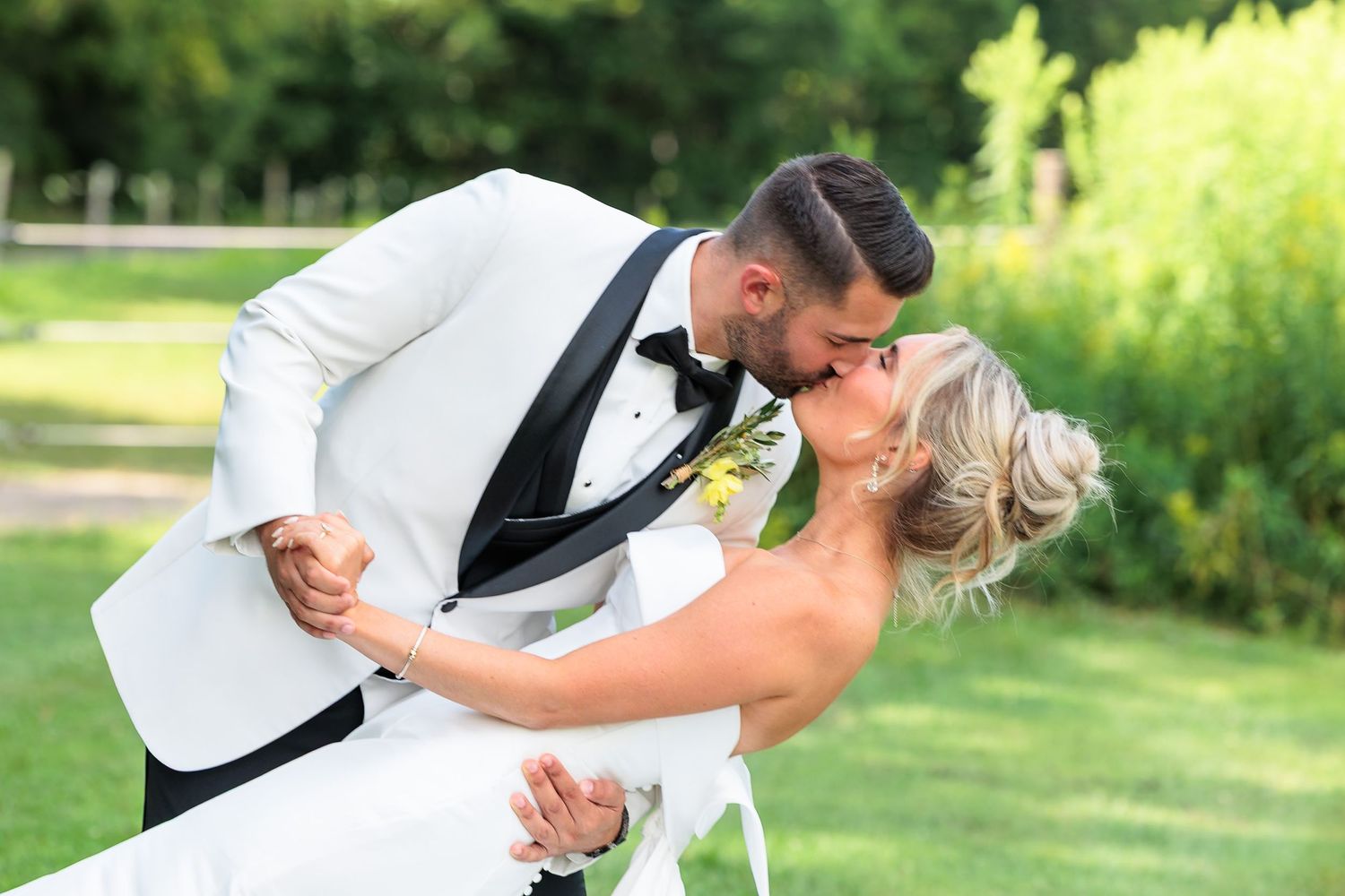 Groom in white tuxedo dipping and kissing bride in white dress on green lawn.