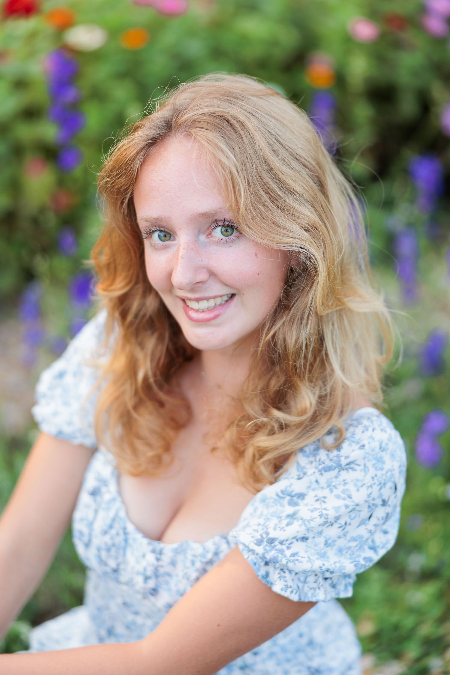 Smiling young woman with curly blonde hair wearing a floral dress, posing outdoors among colorful flowers.
