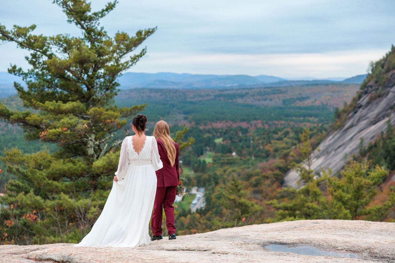 Two brides stand on a rocky overlook admiring a scenic mountain valley view on their wedding day.