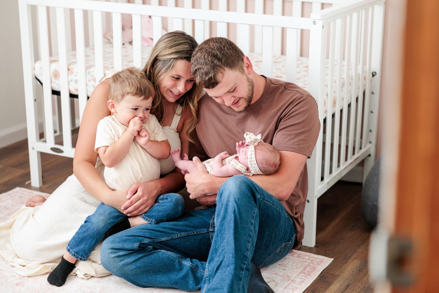 Young family sits on nursery floor by white crib, parents smiling at toddler and newborn baby.