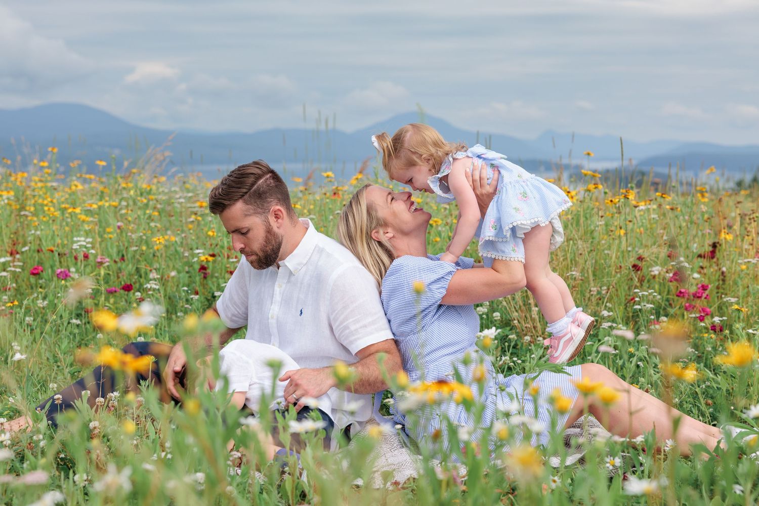 Family sitting in a colorful wildflower field with mountains in the background on a cloudy day.