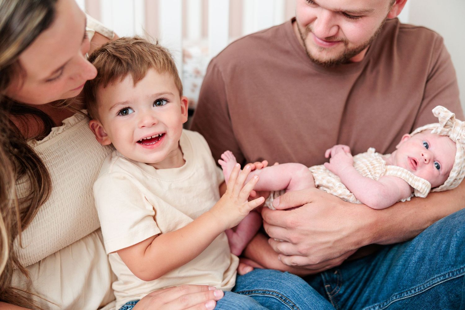 Smiling toddler boy reaching for newborn baby sisters feet while parents look on lovingly.