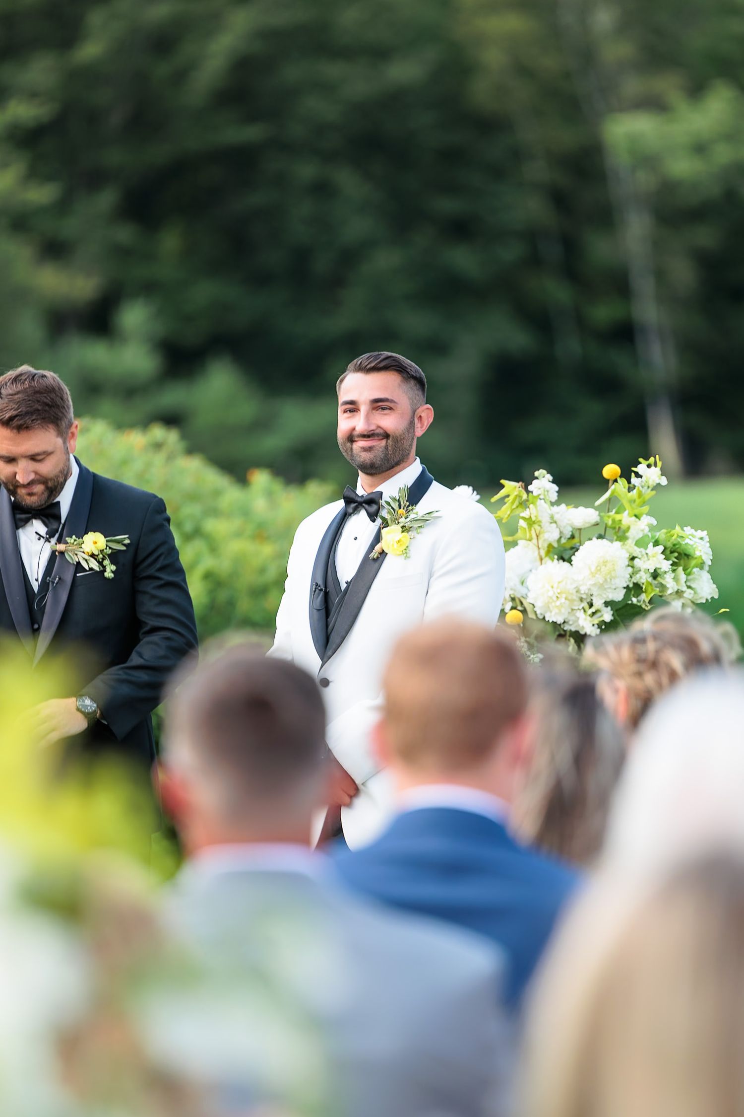 Smiling groom in white tuxedo watching bride walk down the aisle at outdoor garden wedding ceremony.