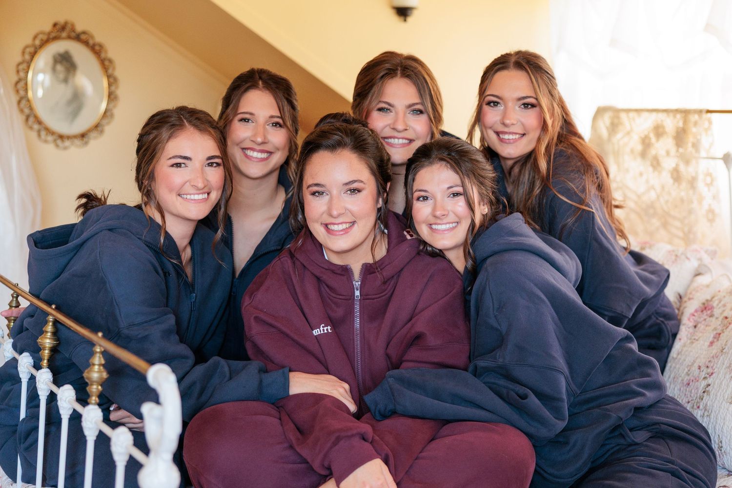 Bride surrounded by six bridesmaids in navy hoodies posing together on staircase before wedding ceremony.