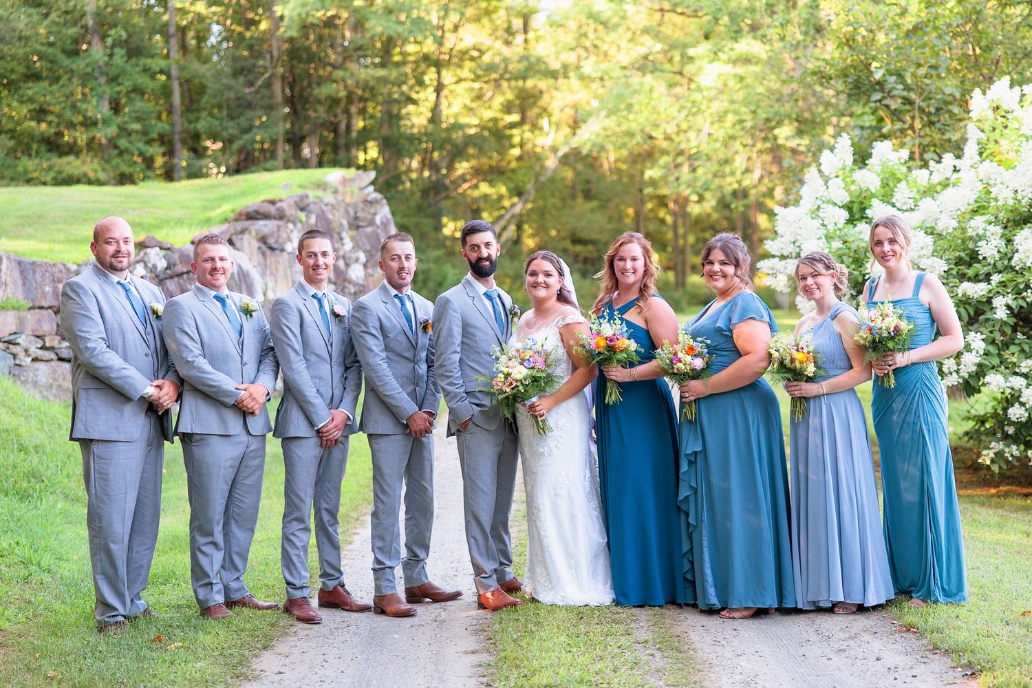 Smiling wedding party stands together on scenic gravel path, featuring blue bridesmaid dresses and gray groomsmen suits.