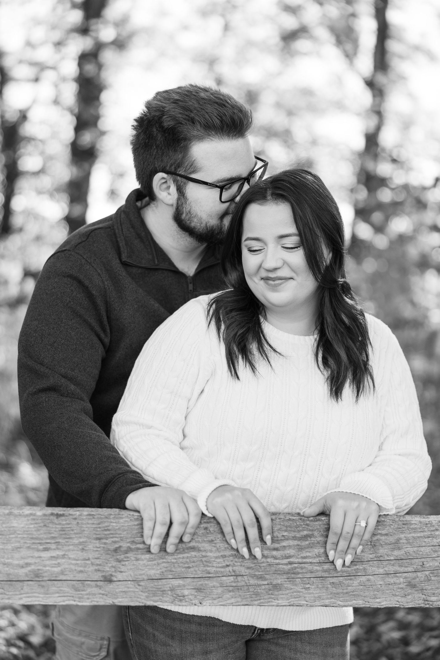 Black and white photo of a couple leaning on a wooden fence, man kissing womans head tenderly.