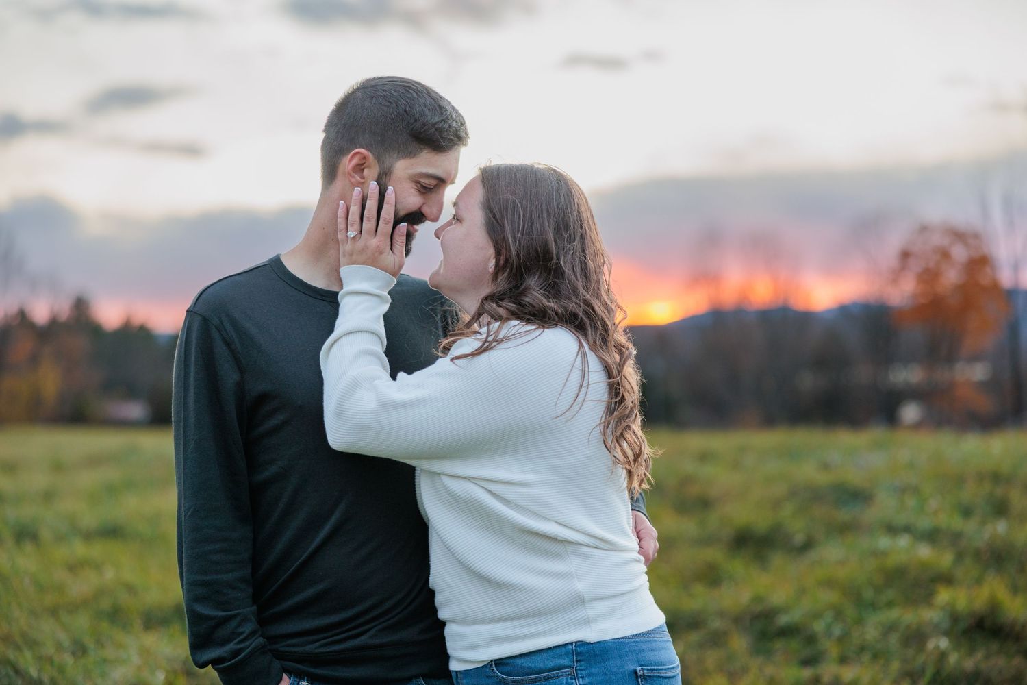 Couple sharing a romantic almost-kiss at sunset in an open field with colorful sky in the background.