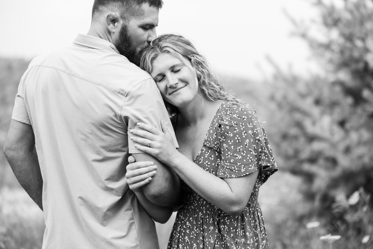 Black and white photo of couple embracing tenderly outdoors with soft natural background lighting.