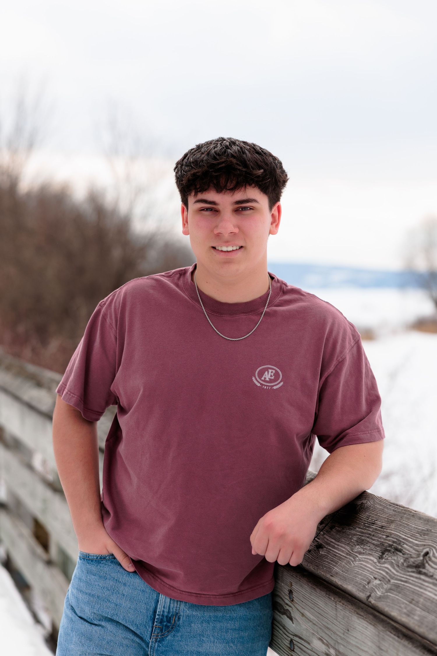 Young man in maroon t-shirt leaning on wooden fence in snowy winter landscape, smiling outdoors.