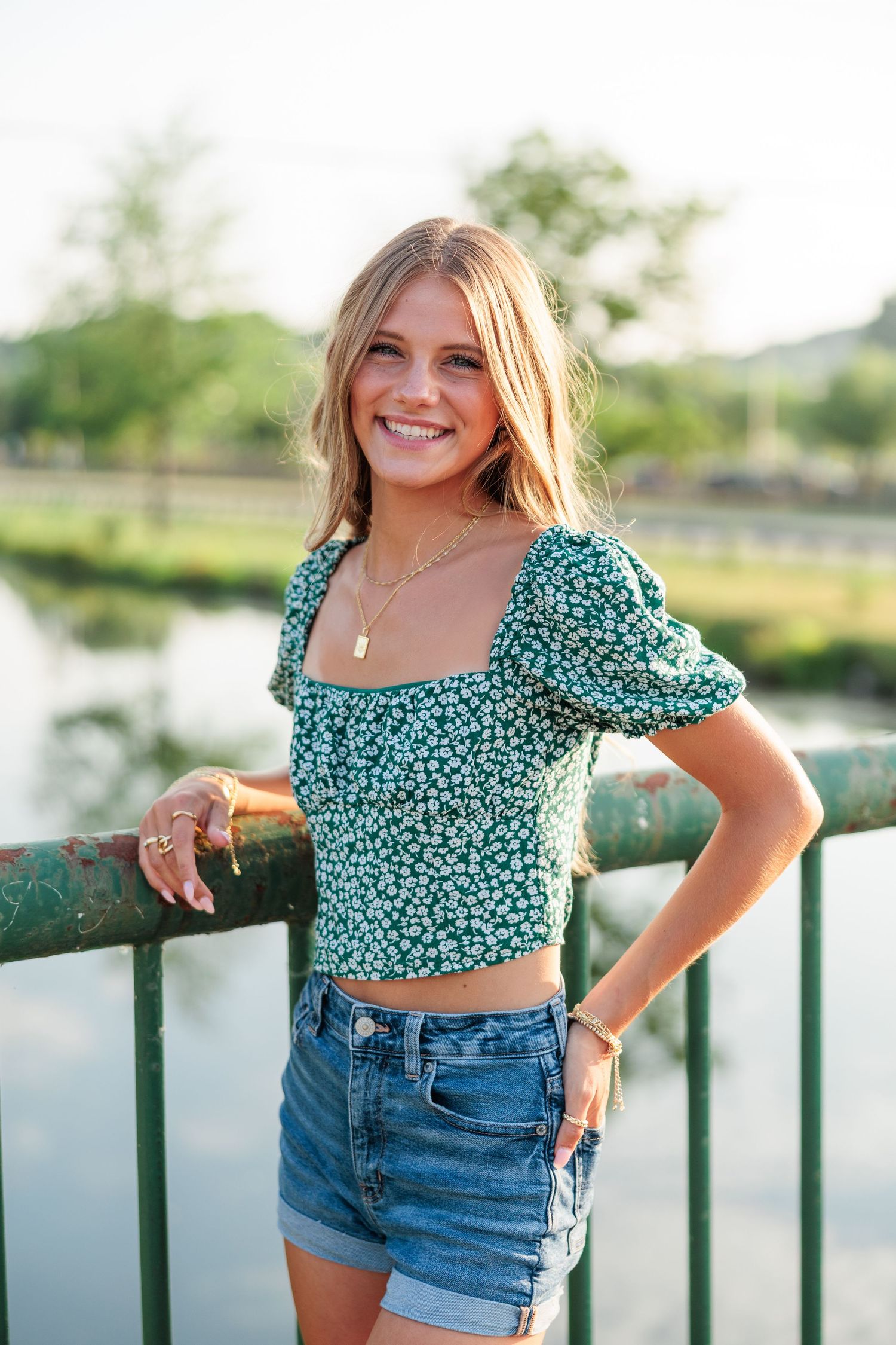 Smiling young woman in green floral crop top and denim shorts leaning on a bridge railing outdoors.