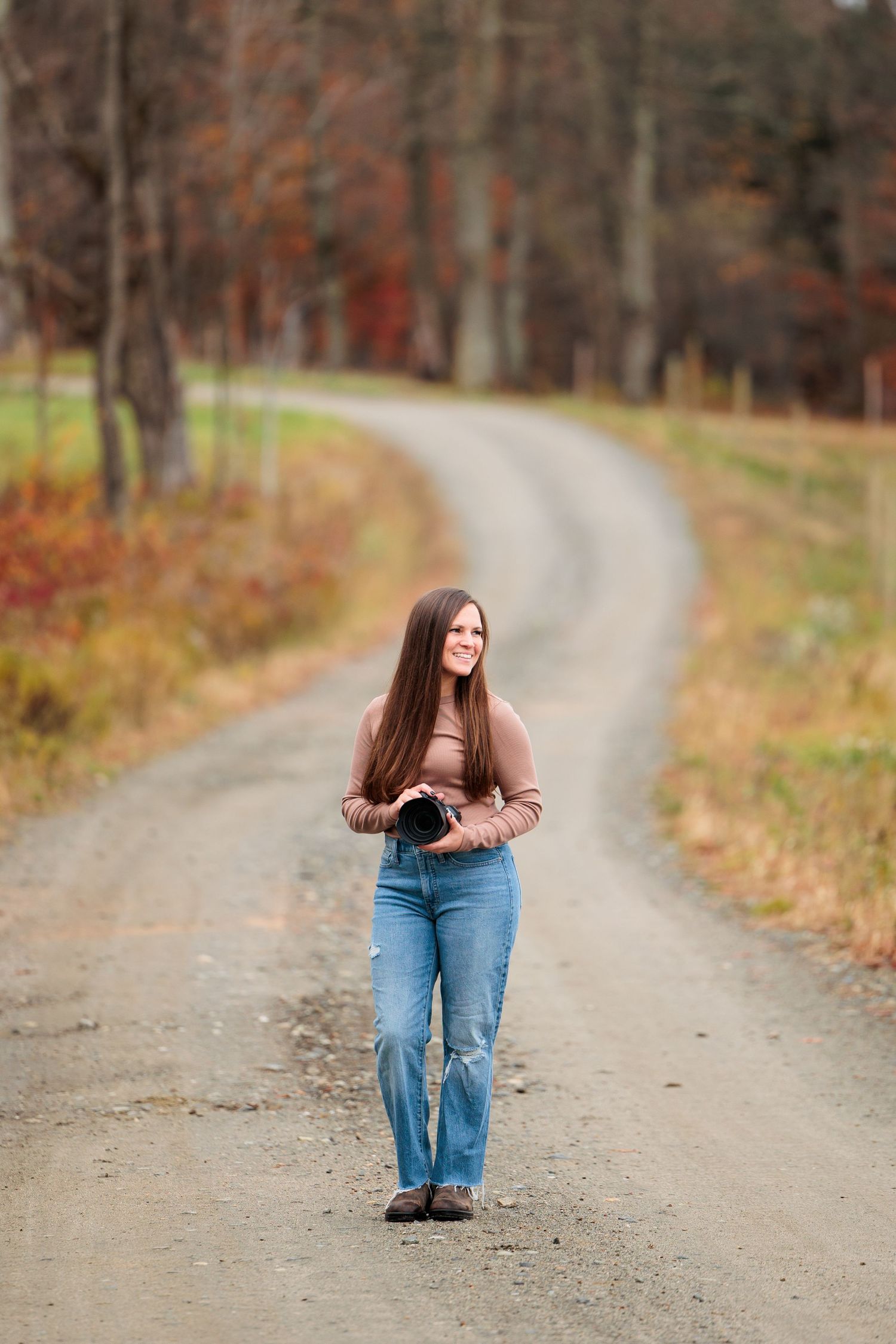 Young woman with long brown hair walking on a rural dirt road surrounded by autumn trees.