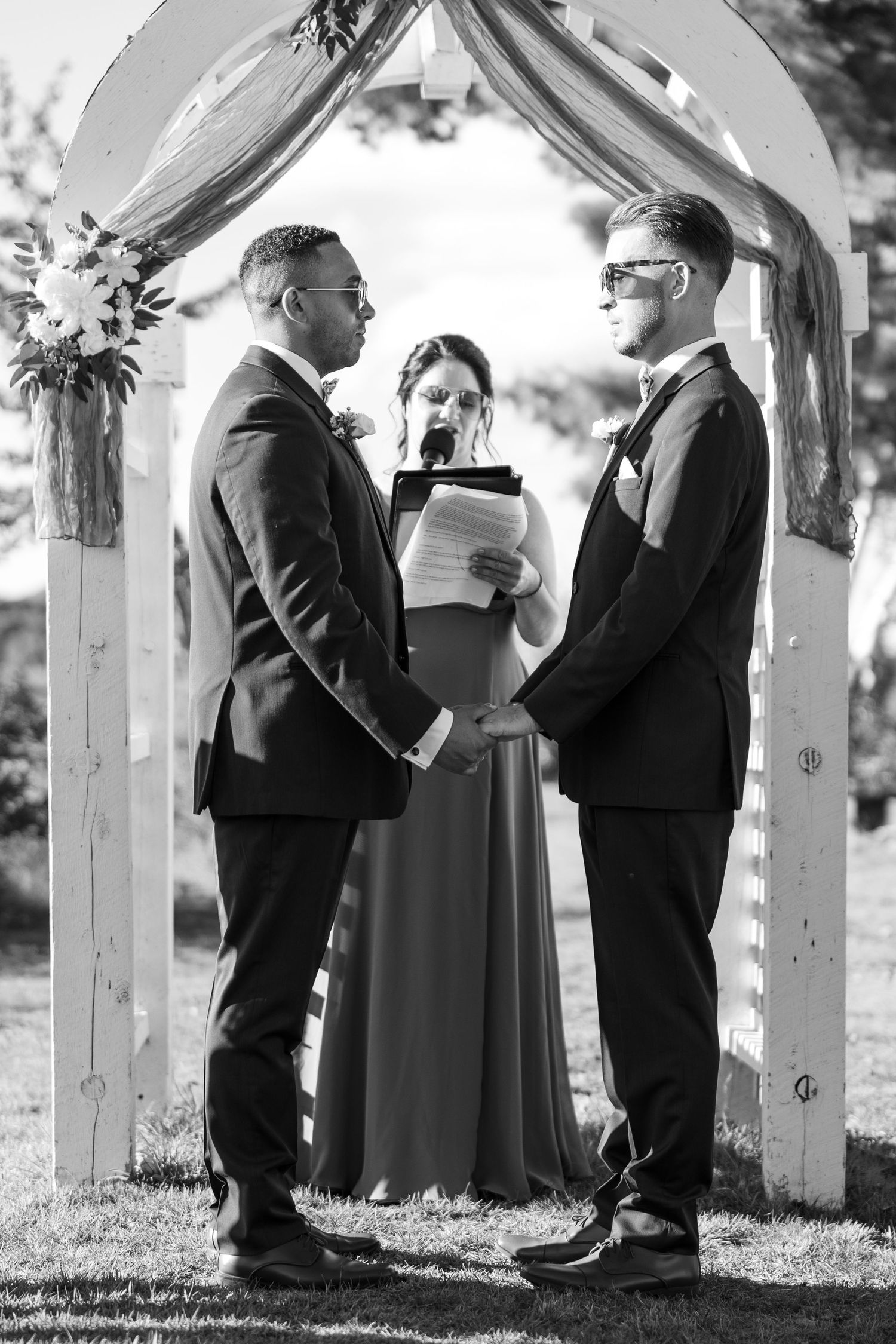 Black and white photo of wedding ceremony participants under white wooden arch.