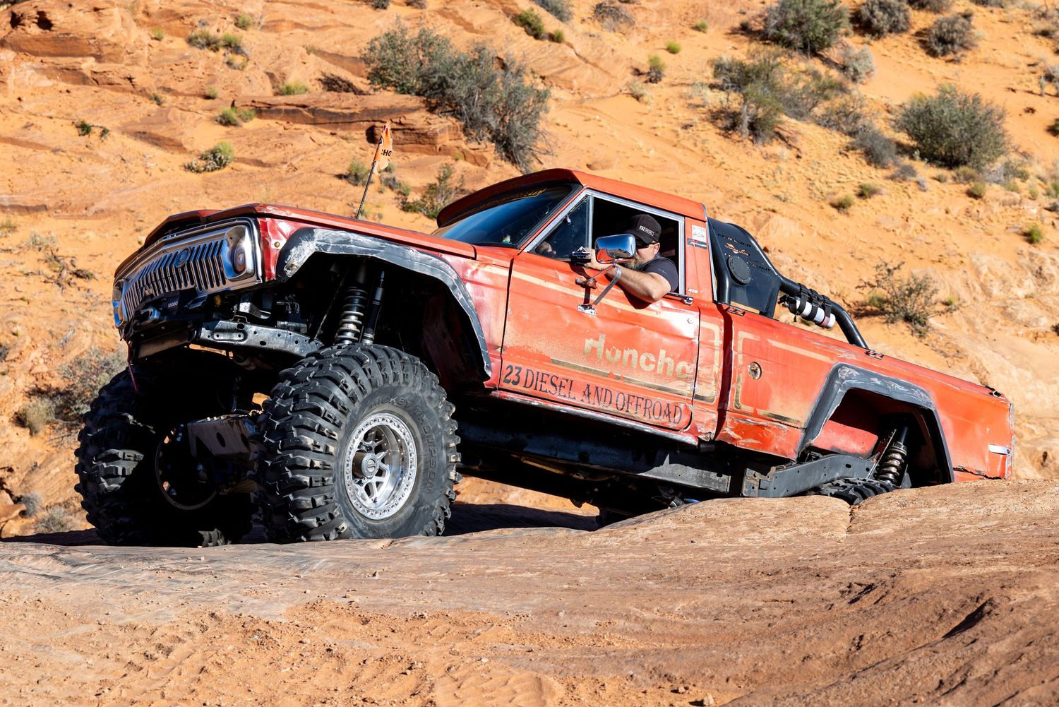 Red modified Jeep truck rock crawling on steep sandstone terrain in desert landscape.