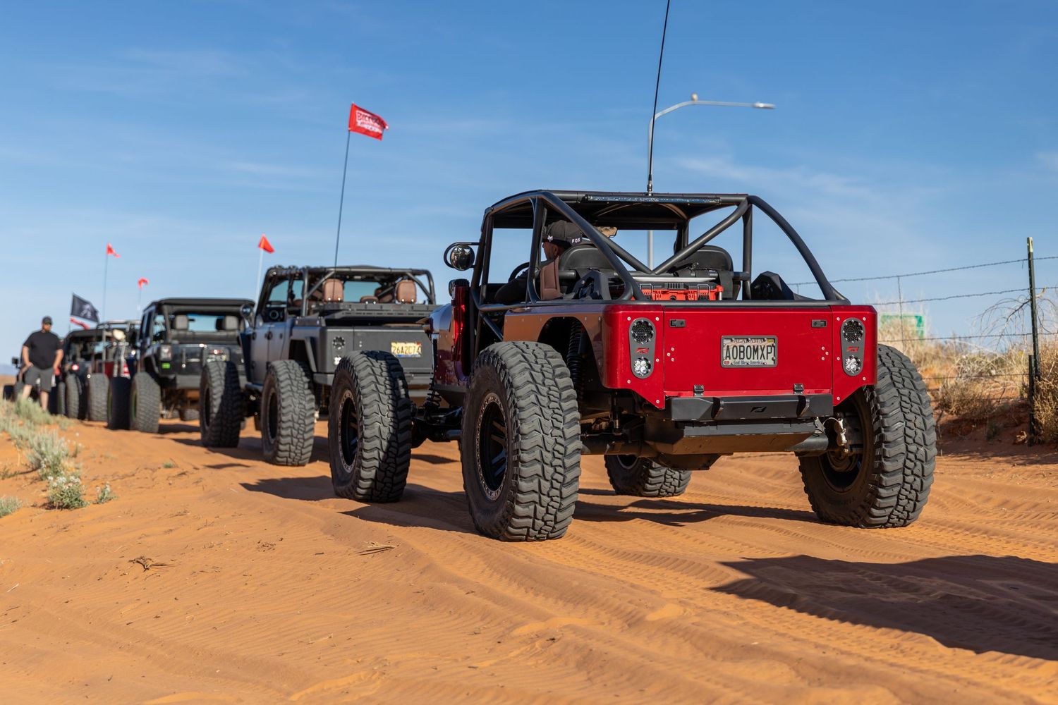 Line of modified off-road Jeeps and buggies parked on red sand trail under blue sky.