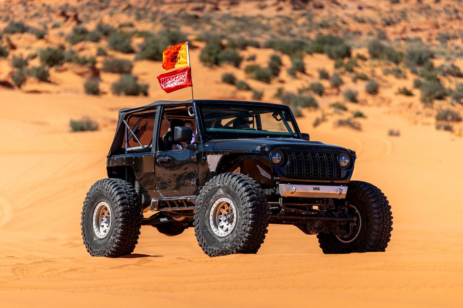Black lifted Jeep Wrangler driving through sandy desert terrain with red and yellow flag.