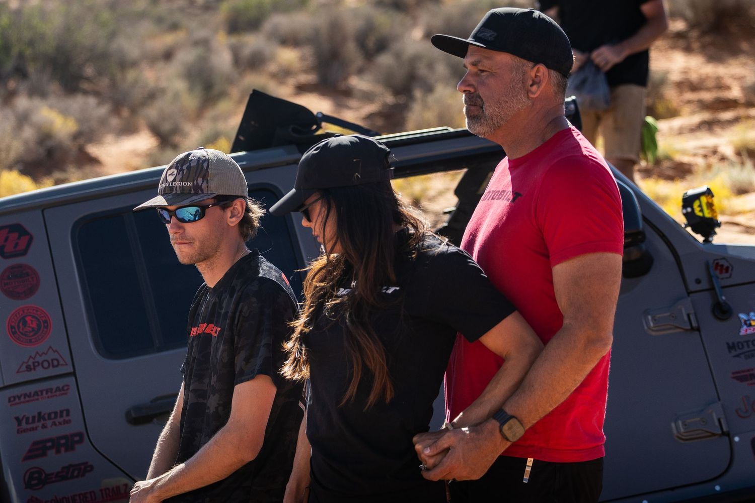Three off-road enthusiasts watching rock crawling event near modified Jeep in desert.