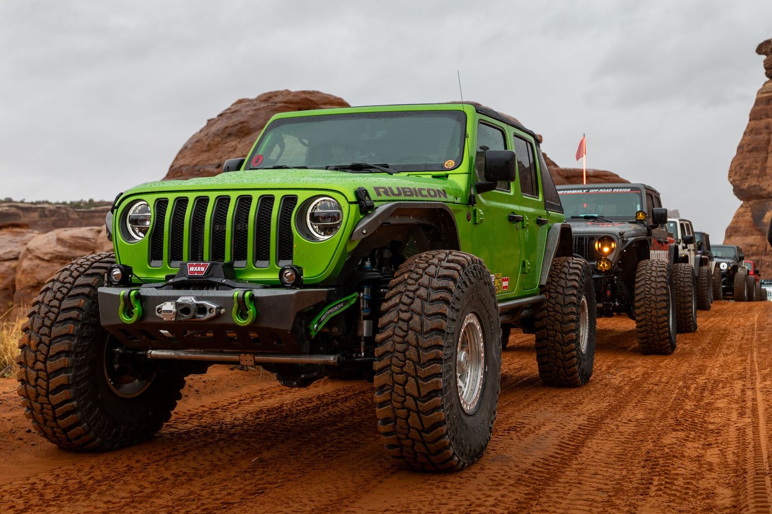 Bright green lifted Jeep Wrangler Rubicon leading a convoy on a rugged red dirt off-road trail in Moab.