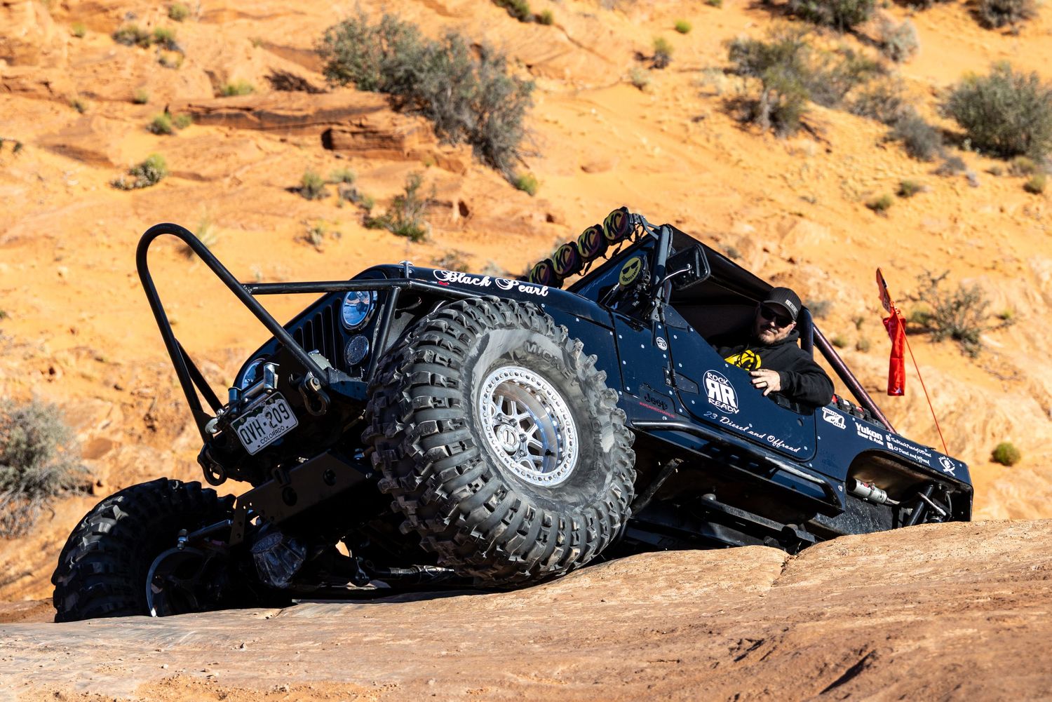 Black lifted off-road Jeep crawling over rocky desert terrain at steep angle with red flag.