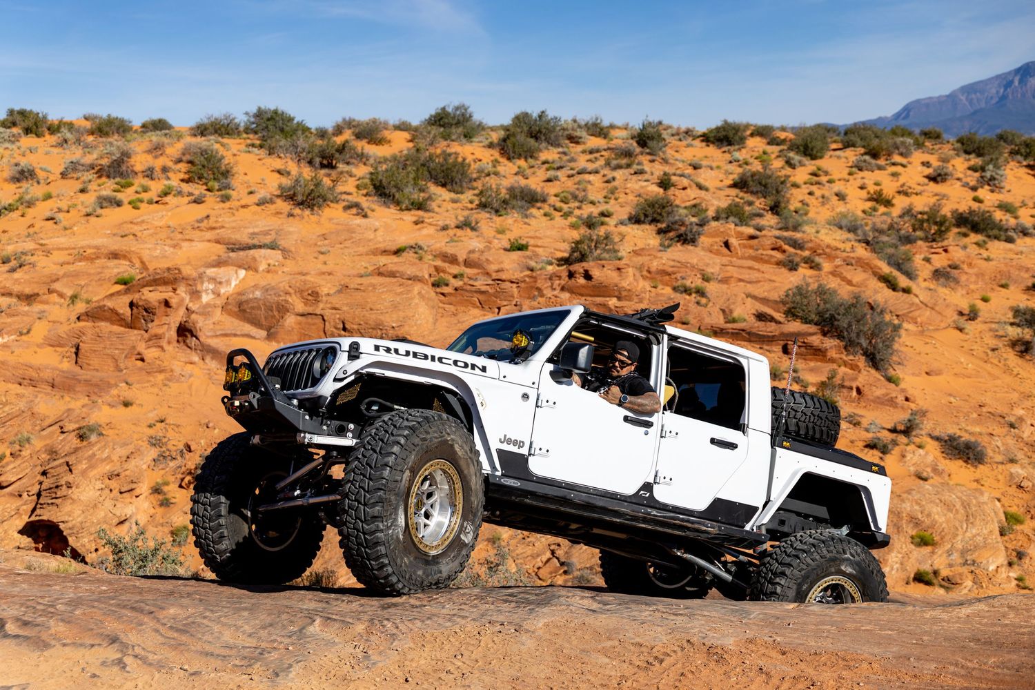 White lifted Jeep Gladiator Rubicon off-roading over rocky desert terrain under blue sky.