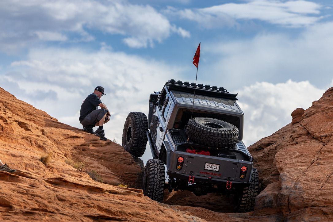 Man crouching beside lifted 4x4 off-road vehicle descending rocky terrain in desert landscape.