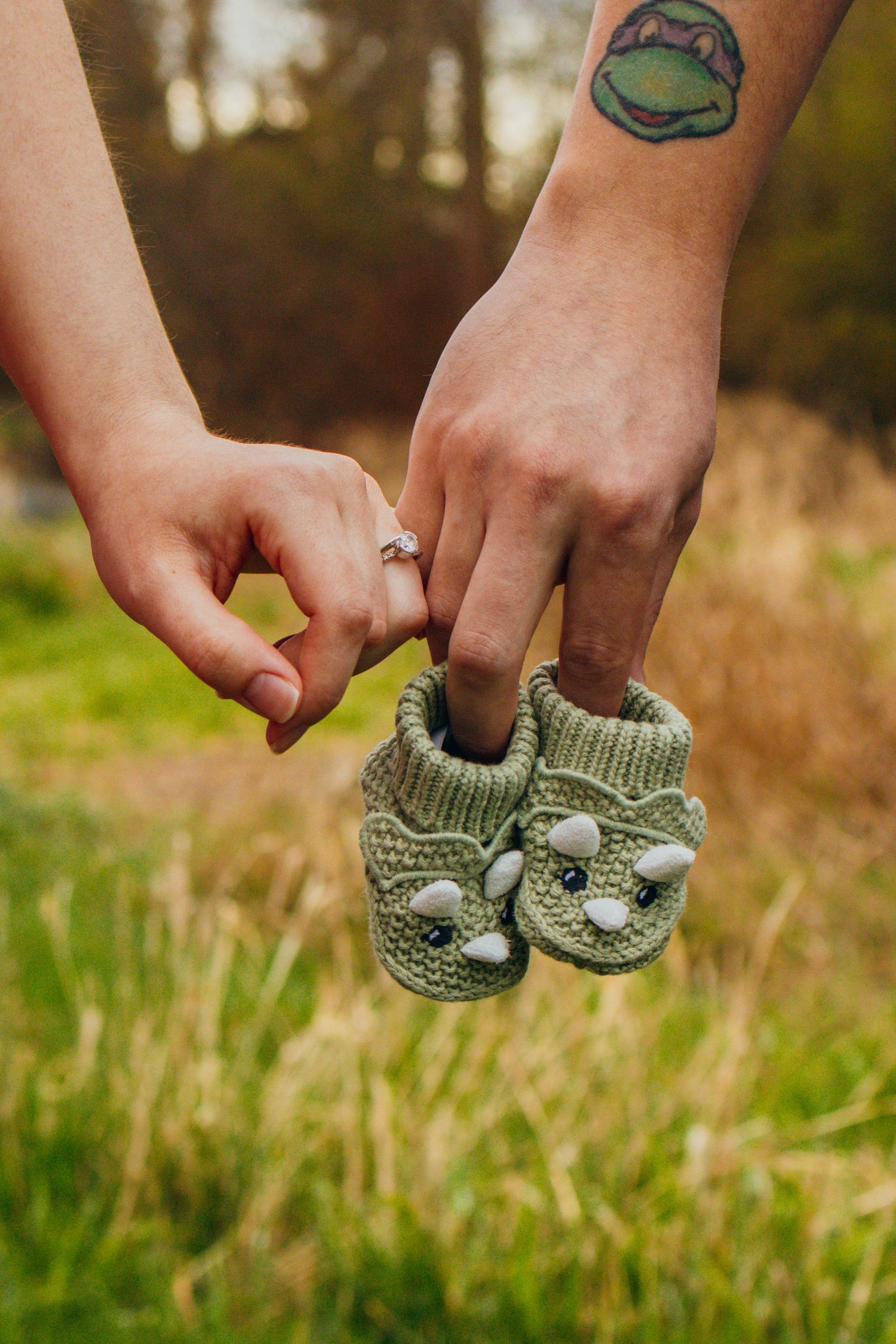 Hands holding baby booties shaped like a dinosaur. They are locked at the pinky fingers on a green grassy back drop at a maternity session.