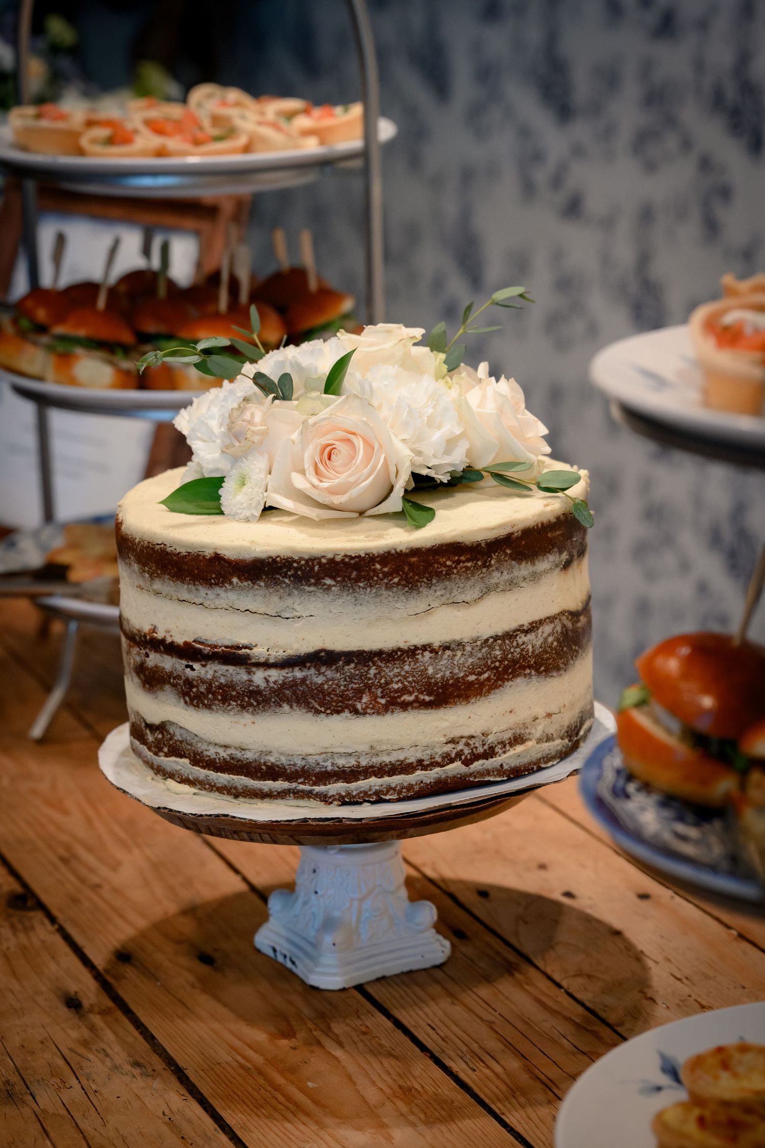 A rustic chocolate layer cake decorated with peach roses and white flowers sits on a wooden table with a vintage cake stand.