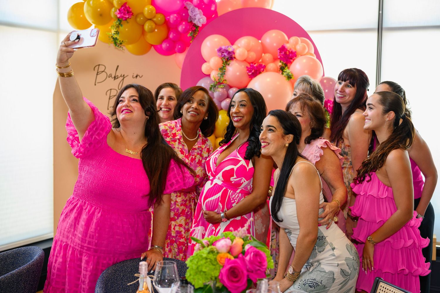 Group of women in pink outfits take a selfie at a colorful baby shower party.