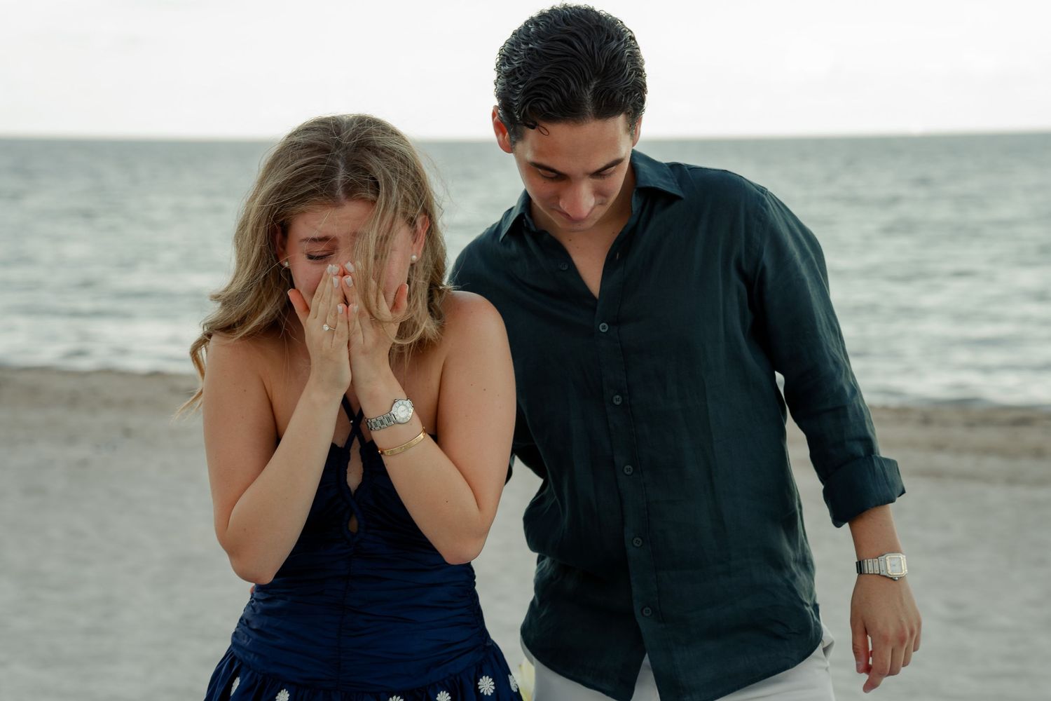 Couple sharing a joyful moment on a sandy beach, woman covering her face with hands, man smiling beside her.