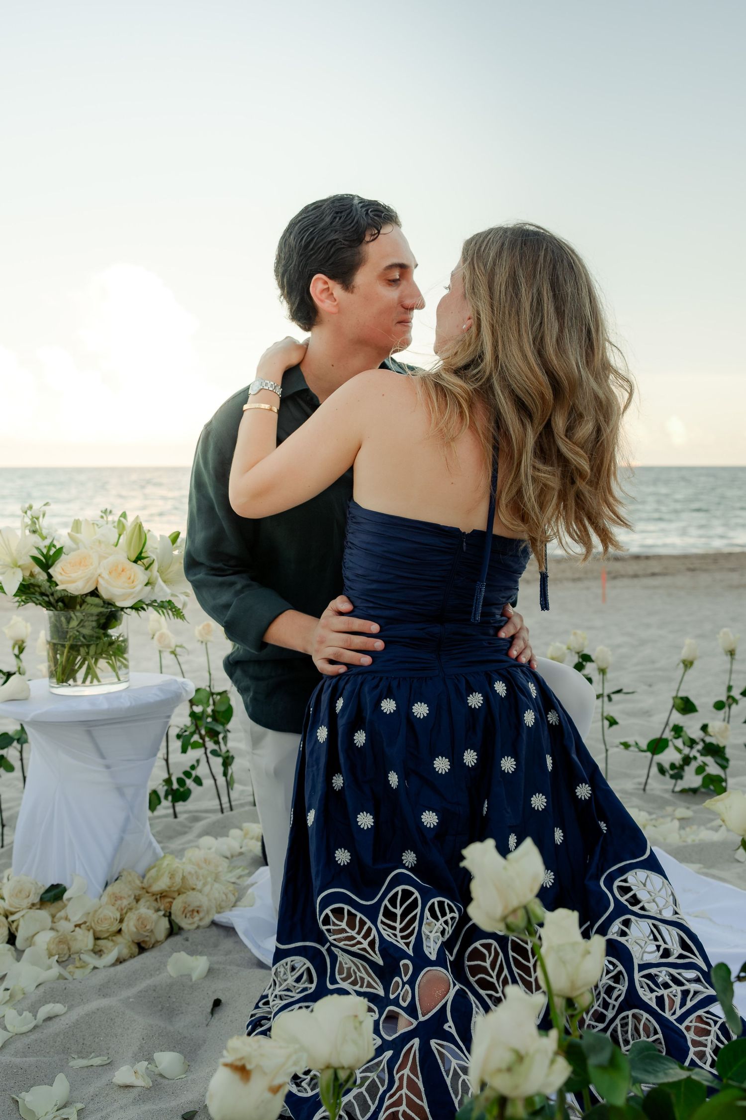 Couple embracing on a romantic beach setup with white roses and ocean backdrop at sunset.