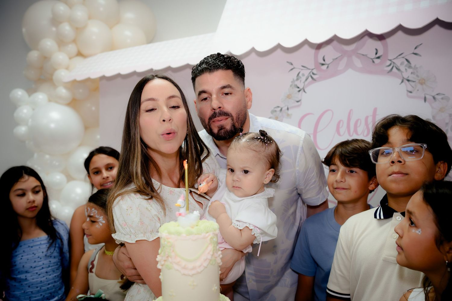 Family celebrating baby’s first birthday with a white cake, balloons, and decorations in a festive party setting.