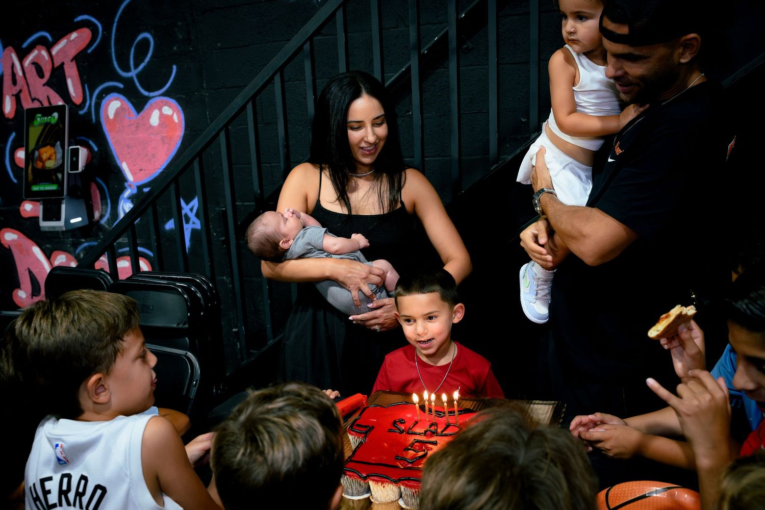 Family gathering with children around a birthday cake with candles, parents holding babies near colorful graffiti wall.