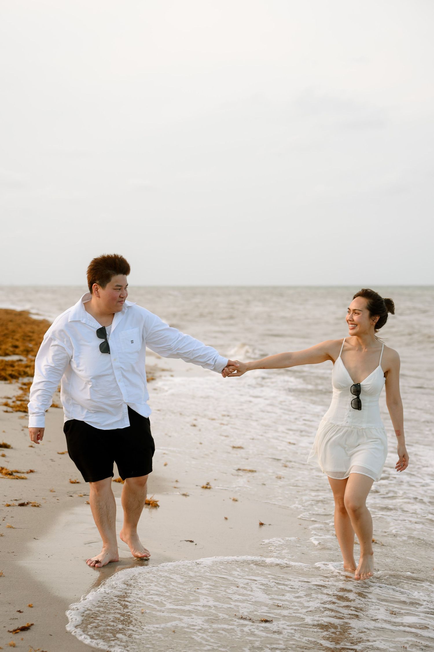 Happy couple holding hands and walking barefoot along a sandy beach shore in matching white outfits.