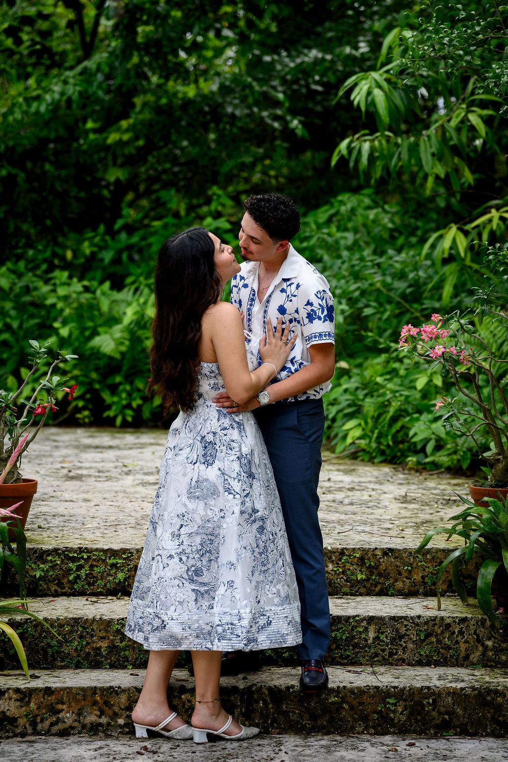 Couple in matching blue floral outfits embracing on stone steps surrounded by lush green tropical garden.