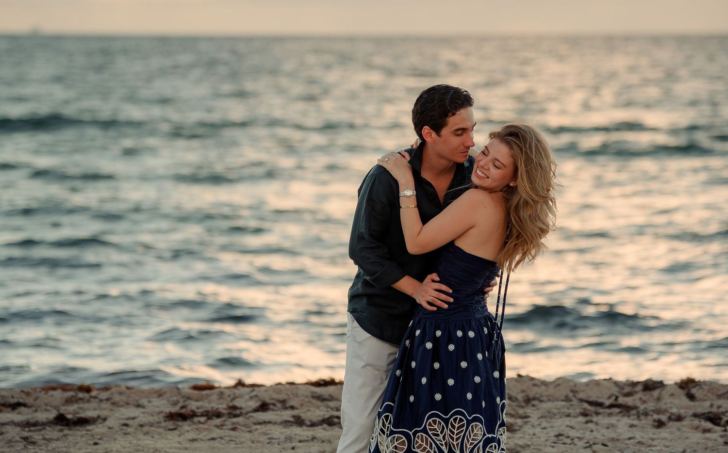 Romantic couple embracing on a sandy beach at sunset, with calm ocean waves in the background.