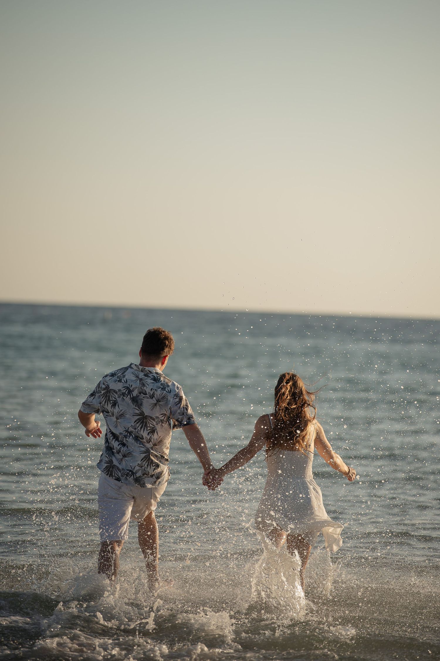 Couple holding hands running into ocean waves on a sunny beach, man in floral shirt and woman in white dress.