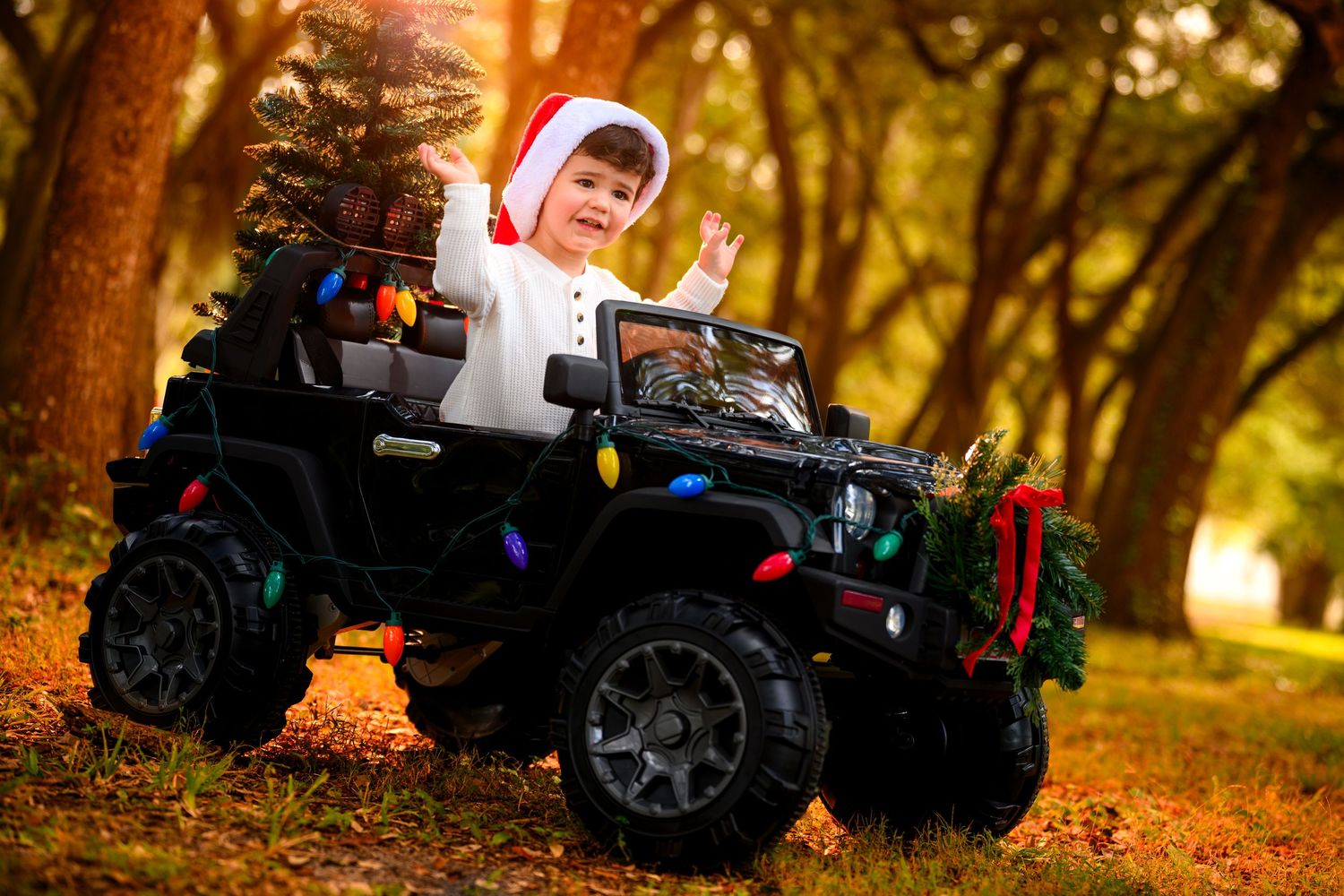 Happy toddler in Santa hat waving from decorated toy jeep in autumn forest.
