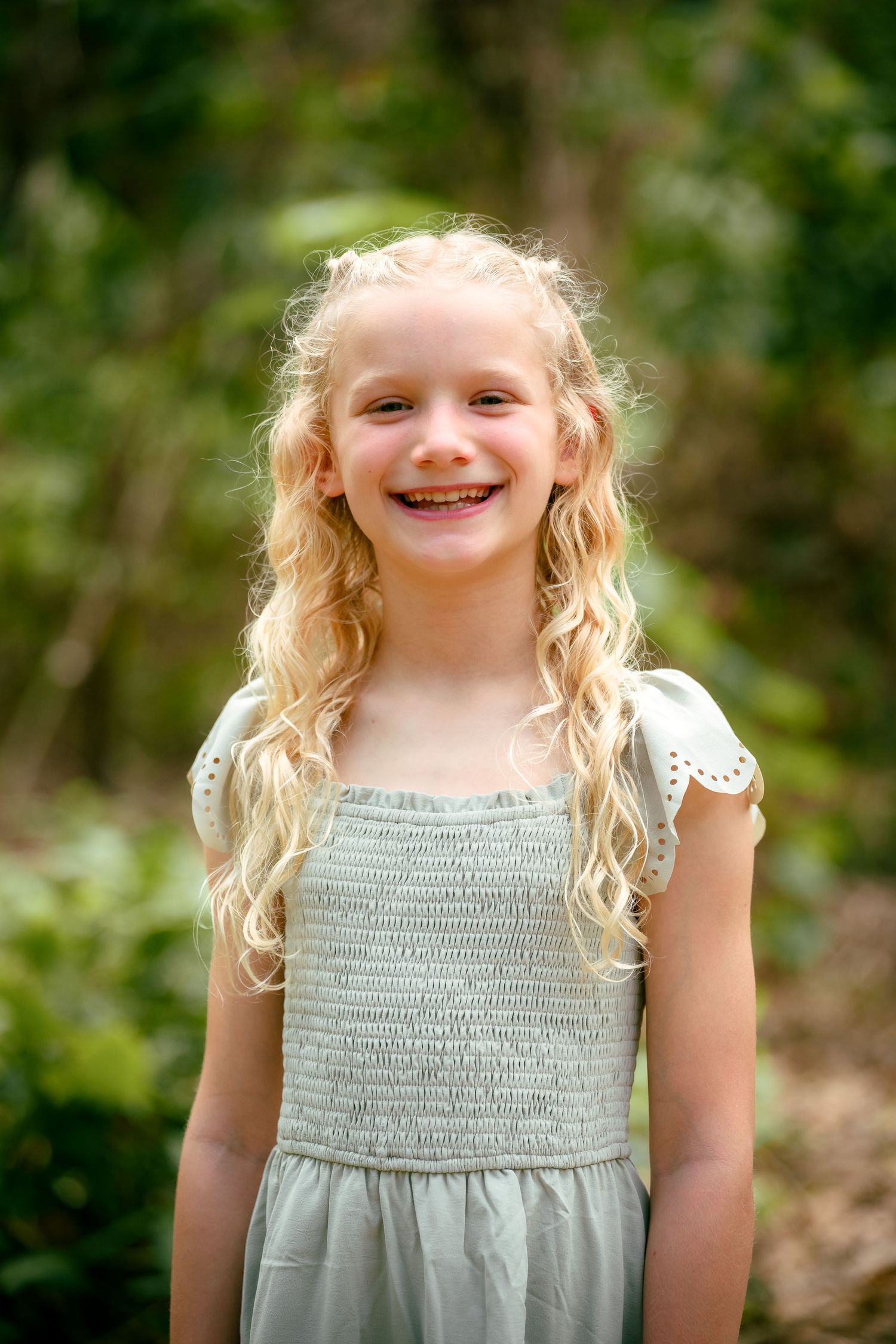 Smiling young girl with curly blonde hair wearing a light gray smocked dress, standing outdoors in a green garden setting.