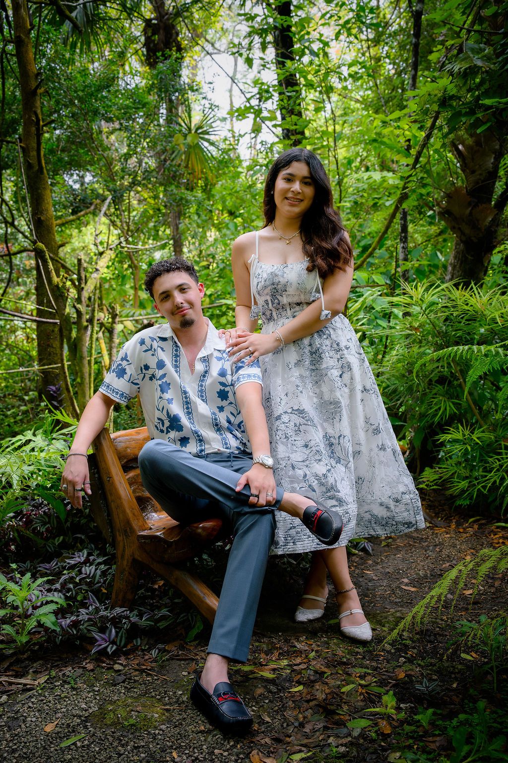 Couple posing in lush green garden, man seated on wooden bench, woman in floral dress standing beside him.