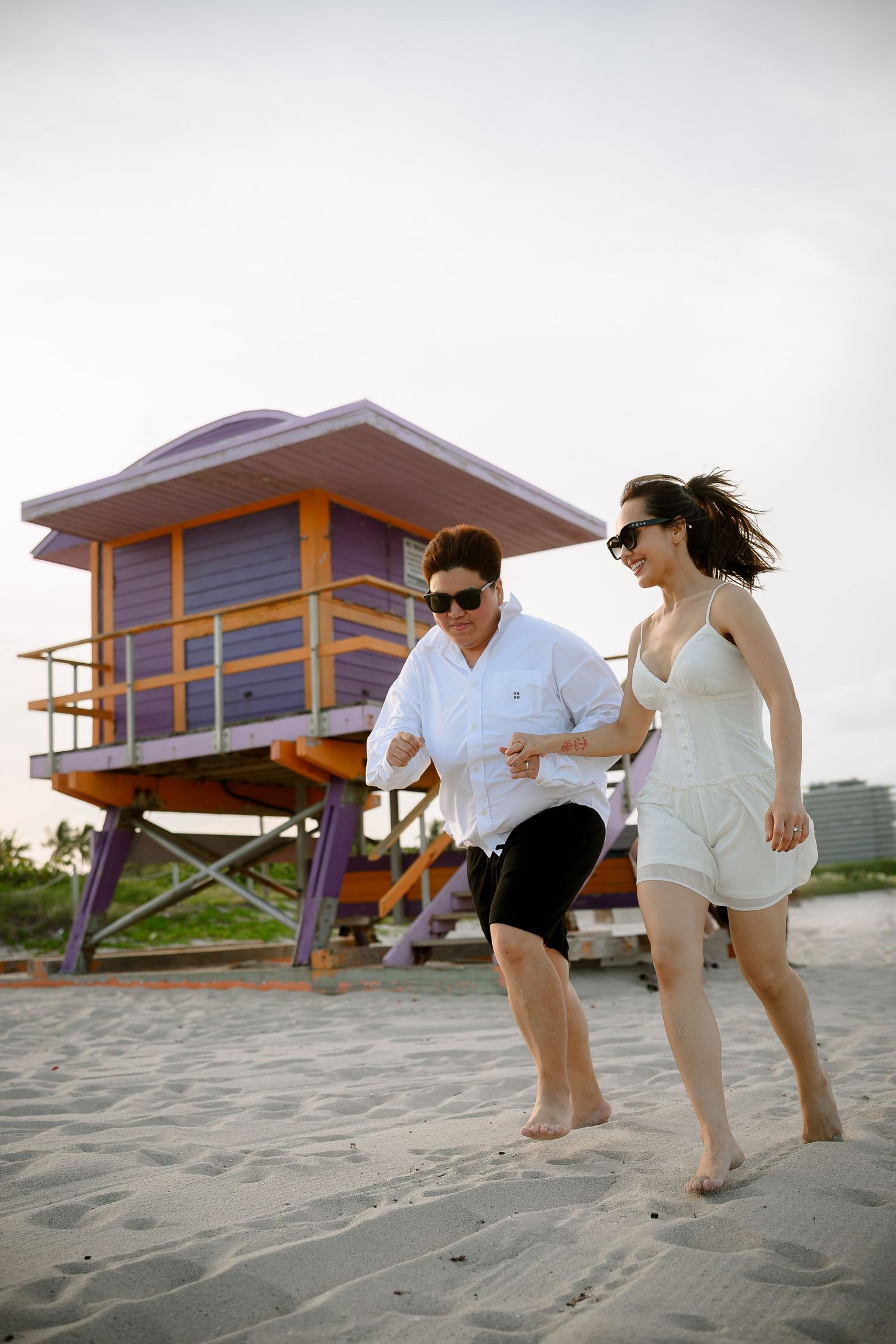 Couple running and laughing on Miami Beach sand near a colorful purple and orange lifeguard stand.