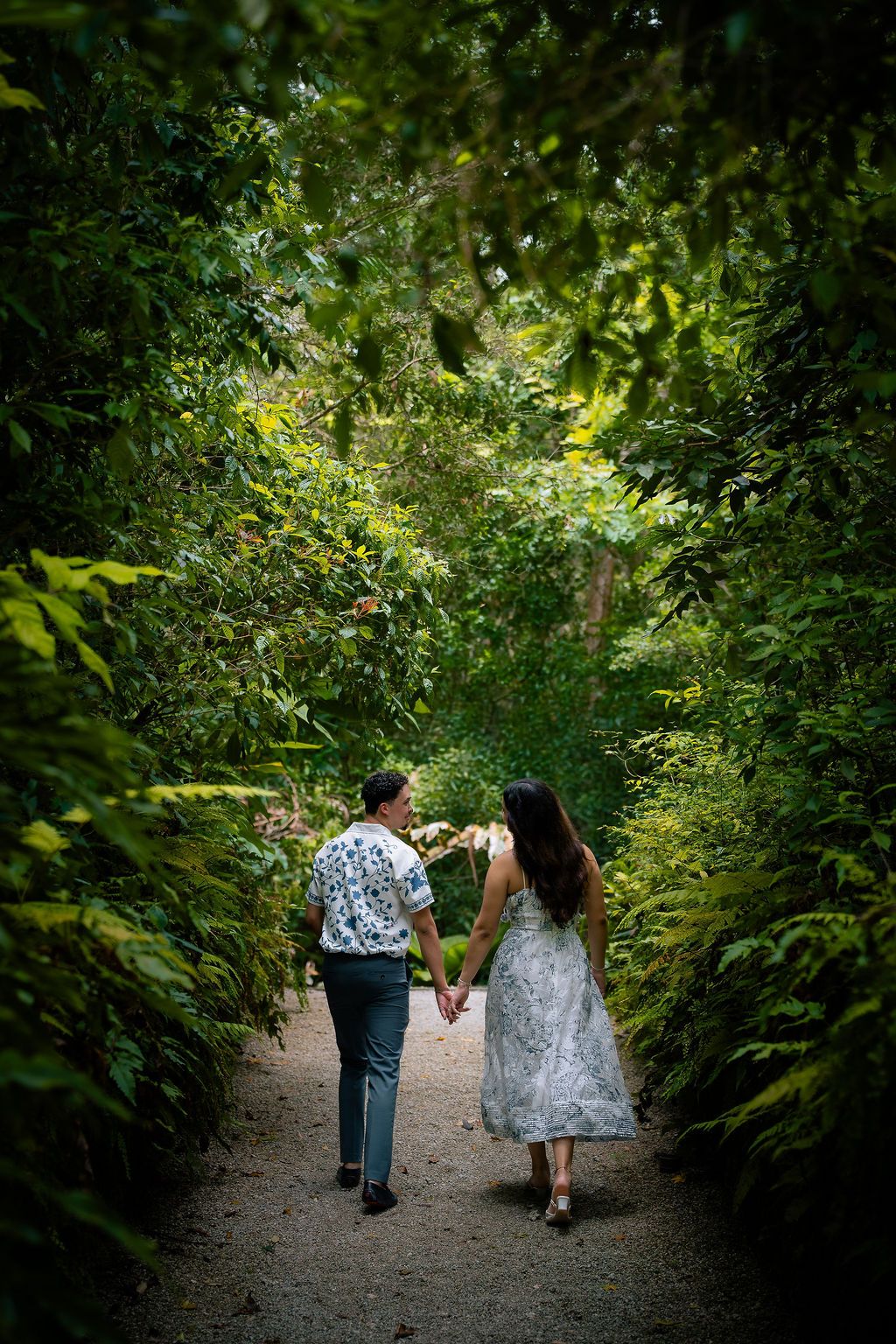 Couple holding hands walking down a lush green garden path surrounded by tropical foliage.