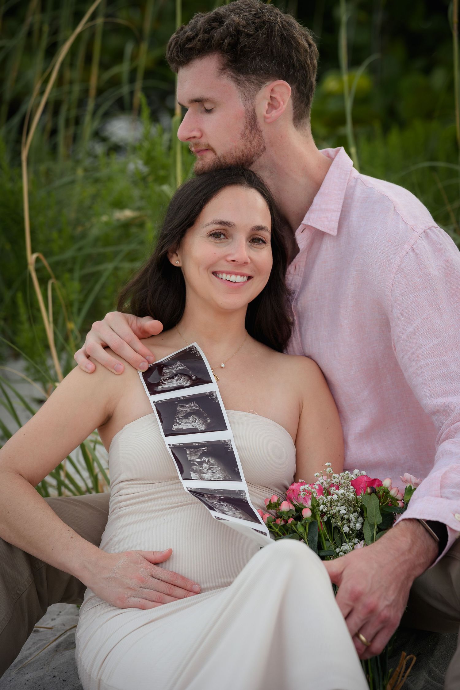 Smiling pregnant woman in white dress holds ultrasound photos and flowers while partner kisses her head outdoors.