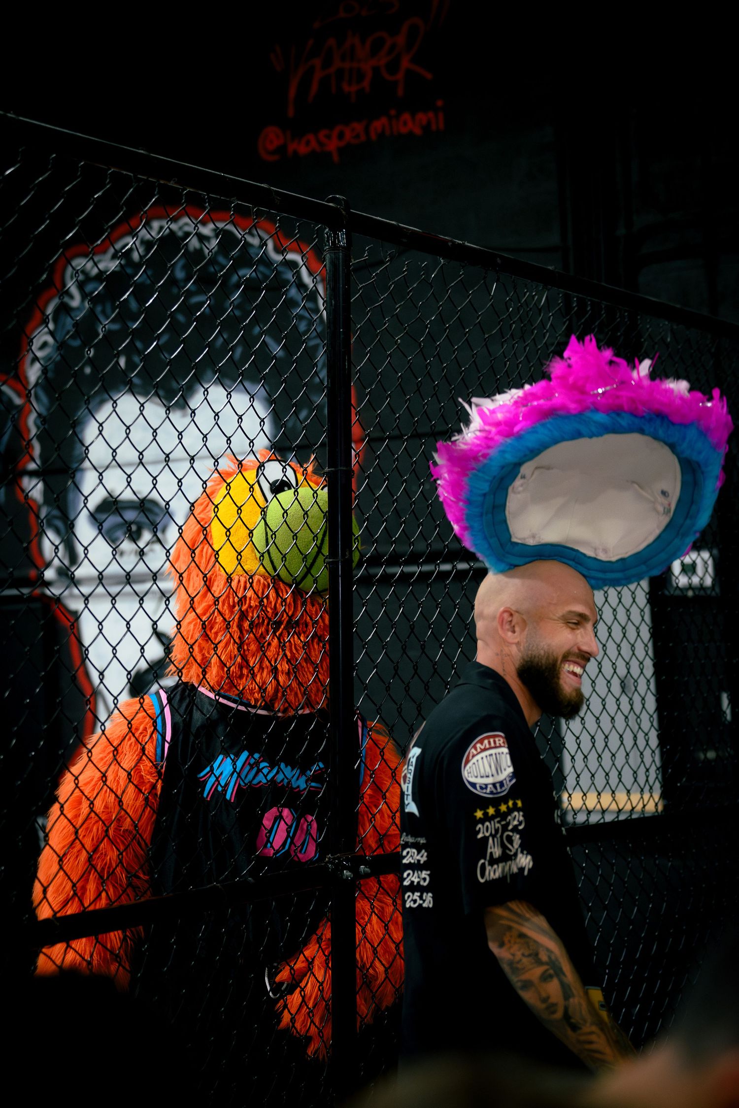 Man with colorful hat smiles near Miami Heat mural and chain-link fence at street art event.