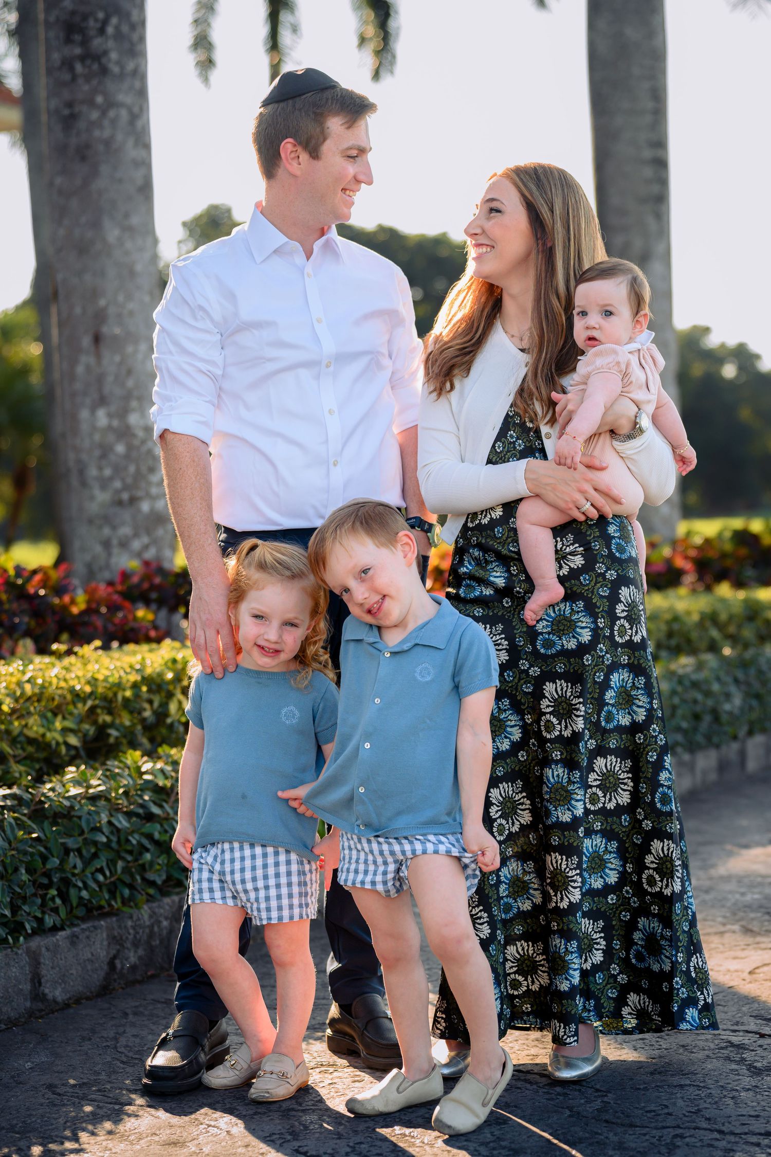 A happy family of five poses outdoors among palm trees, with parents and three young children dressed in coordinating outfits.